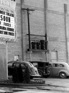 A black and white photo.  On the side of the building, three large power transformers sit on top of a beam supported by two poles. One pole extends higher, allowing four lines to extend over the parking lot. Three vintage cars are parked beside the building. On the left edge, a portion of the theater's sign is visible. - , Utah