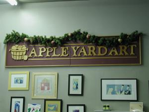 Mounted on the wall is a sign reading, 'Apple Yard Art,' with the image of a barrel full of apples.  Below the sign are several small framed art pieces. - , Utah