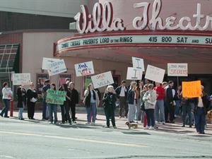 A couple dozen people stand on the sidewalk in front of the Villa, displaying signs supporting the theater.