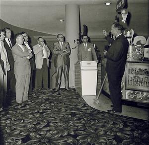 Several men in business suits watch as a man speaks while standing near the south end of the concession stand.  A stairway to the lower lobby is in the background on the left. A section of the original planter box is visible in the middle. - , Utah