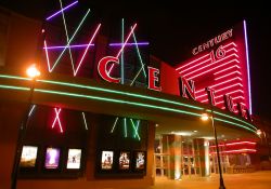 Neon on the front of the theater at night.  On the left side of the entrance are six poster cases, with flashing multi-colored lights above them.  On the right side is an outside ticket window and a tall tower bearing the name 'Century 16'.