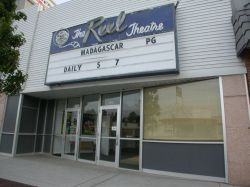 The sign for 'The Reel Theatre' is above the entrance and has a four-line attraction board.  The entrance is a diagonal wall with windows and two doors.  On the left of the entrance there may have been an ticket booth.
