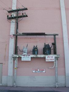 A color photo of three transformers mounted on a beam between two poles. The brick behind them is painted pink. Sign read, 'Park at Your Own Risk,' 'Apple Yard, Villa Parking,' and 'Fire Zone No Parking.' - , Utah