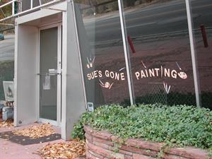 The Apple Yard storefront, viewed at an angle, with the entrance in the background on the left and a planter box with ivy in the foreground on the right.  On the large glass window panes, angled outward toward the top, are the words, 'Sue's Gone Painting.' - , Utah