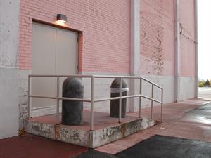 Two exterior doors with no handles open onto a concrete platform with a metal railing. A short ramp leads down to a concrete sidewalk. The walls of the building are pink brick with occasional white concrete pillars. A bit of the parking lot is visible in the background. - , Utah
