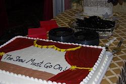 On a table is a cake decorated as stage with the curtains parted to reveal the words, 'The Show Must Go On!'