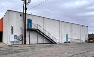 Looking across a parking area at the white brick wall of a building. At the left edge are five electrical panels and a utility pole. A metal staircase with a fenced rail rises to a second-floor door. In the middle of the wall is a single door, with two steps on one side and a ramp on the other.  At the end is a garbage dumpster and a double-door exit. - , Utah