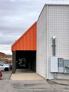 Four large electrical boxes are mounted on a corner wall of white brick building.  Around the corner is a wide sidewalk that leads to a glass door with the number 3 overhead.  Above the sidewalk is sloped roof with orange metal siding. - , Utah