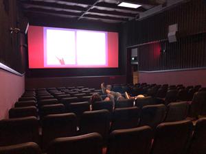 Theater 2 has a center aisle with six seats on either side. In the right wall, just before the screen, is an open doorway with an exit door visible beyond. Three moviegoers sit in the auditorium, one with feet resting on the seat in front. - , Utah