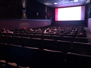 Looking across a long auditorium from the back right corner.  Pre-showtime content displays on the screen while a couple dozen moviegoers wait in their seats.