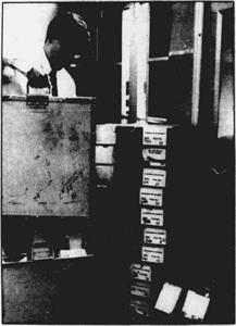 Black and white photograph of a man holding a metal cannister.  A dozen other canisters are stacked on the floor nearby. - , Utah
