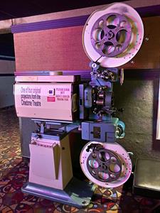 A movie projector stands against a wall, with 35mm film threaded through from a reel on the top to another reel on the bottom. - , Utah