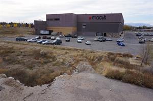 A three-story building sits in the background. Its dark color scheme is contrasted by original brick near a loading area probably originally hidden by the mall.  An open field separates the building from asphalt and broken rebar in the foreground. - , Utah