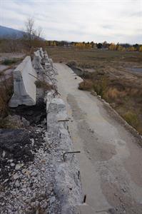 A broken concrete wall with rebar sticking out of the top is protected by a line of a moveable concrete barriers. Below, is a strip of former parking area, with more broken concrete and rebar before dropping down to a field of weeds further down. - , Utah