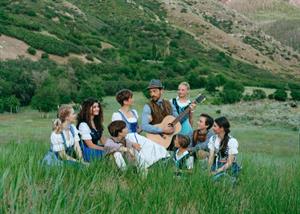 A man with a guitar sits in wild grass, surrounded by seven children and a woman. A mountain rises in the background. - , Utah