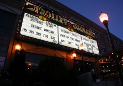 The sign of the Trolley Corners Theatres at night.  The words 'Trolley Corners' appear at the top of the sign and 'Theatres' is written on either end.  Below the name of the theater were sections for each theater with seven lines of copy.